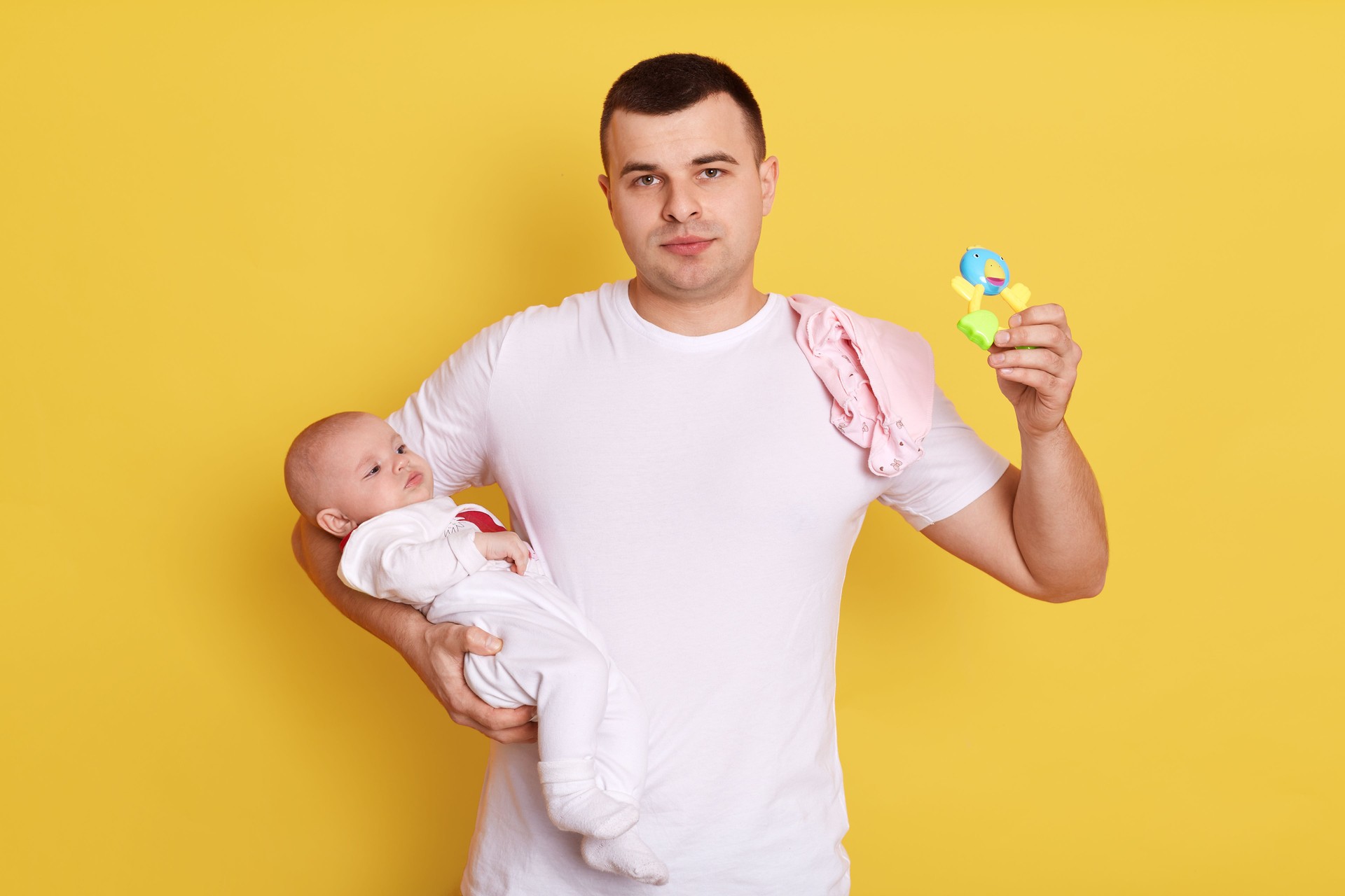 Handsome father and baby child posing isolated over yellow background, dad holds beanbag in hands, wearing casual white t shirt, european daddy newborn daughter.