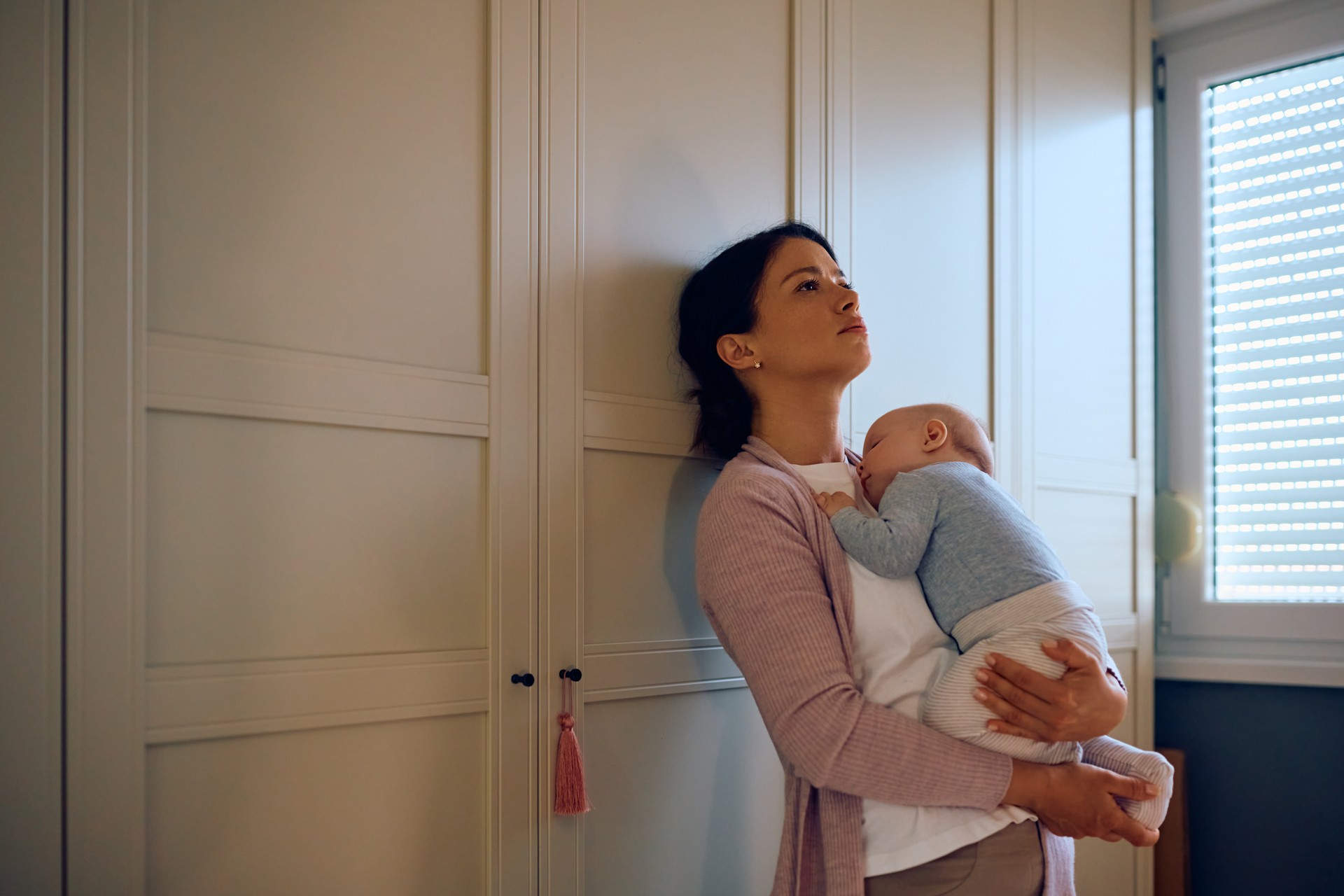 Young sad mother holding her sleeping baby in her arms at home.